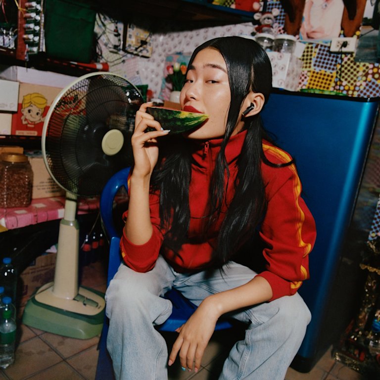 Woman in a red jacket eating watermelon and wearing Marshall Minor IV Black true wireless earbuds in a colorful room.