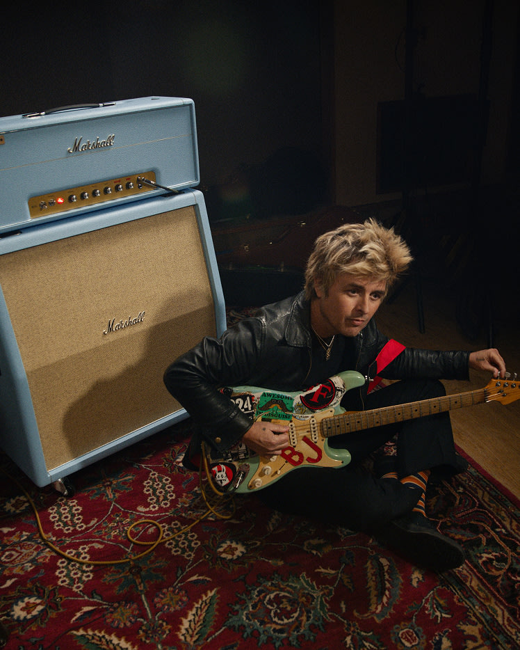Billie Joe Armstrong sits on a patterned rug, playing an electric guitar next to a Marshall 1959 BJA Artist Signature amp stack in a dimly lit room.