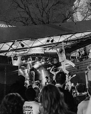 A band performs on an outdoor stage at night under "Mohawk" signage, with an audience watching and one person mid-jump.