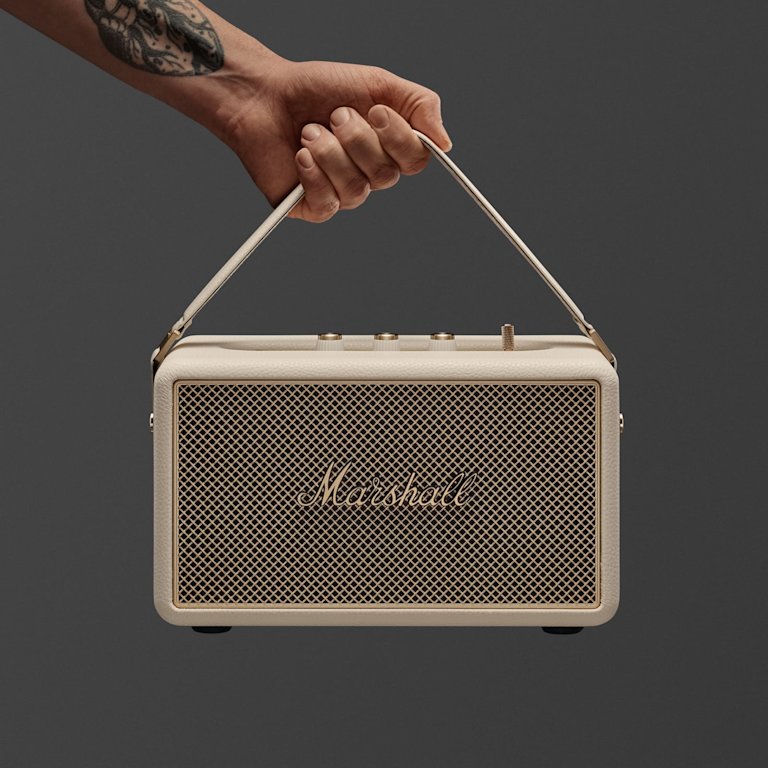 A hand holds a Marshall Kilburn III Cream portable speaker by its strap against a plain green background.