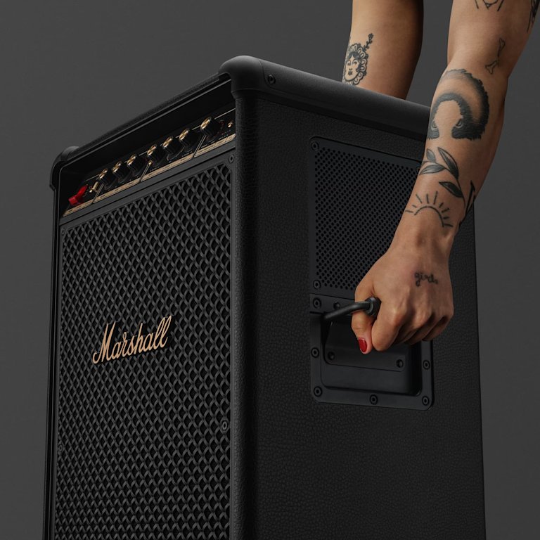 A tattooed arm grips the handle of a large black Marshall Bromley 750 speaker amplifier.