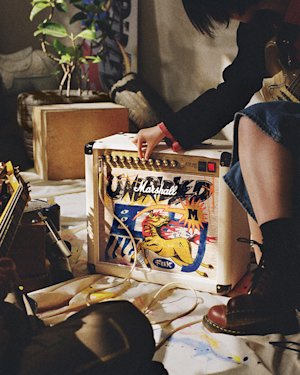 A person adjusts Studio 900 Combo Lunar New Year Edition 2026, sitting on a blanket-covered floor with scattered objects and holding a guitar.