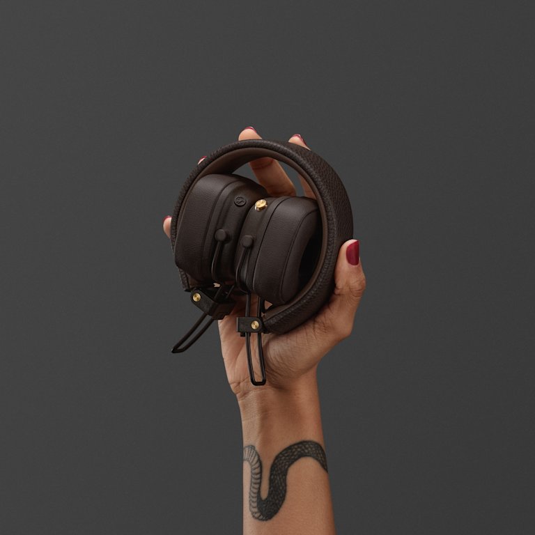 A woman with red nail polish holds Marshall Major V Brown on-ear headphones against a dark gray background.