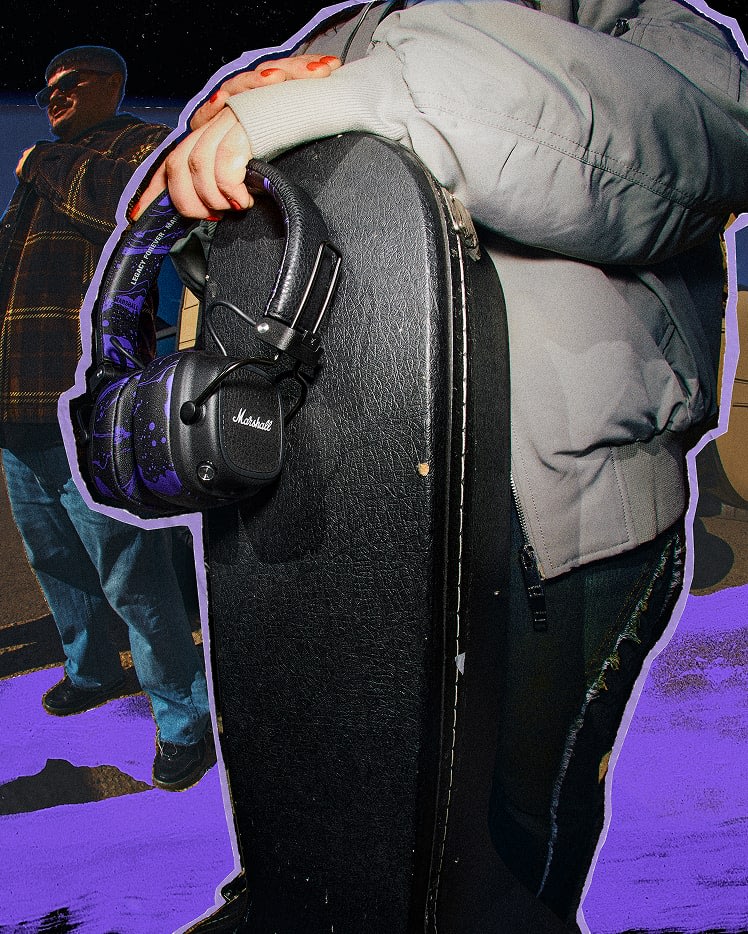 A person in a gray jacket holds a guitar case and Marshall Major V Hendrix Edition headphones.