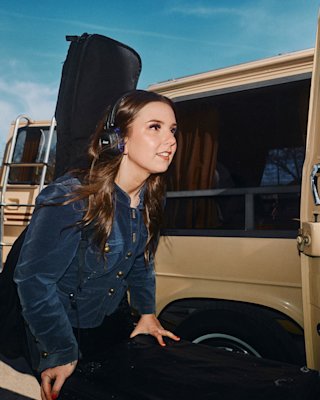 A woman wearing Major V Hendrix Edition headphones carries a guitar case while standing next to a beige van.