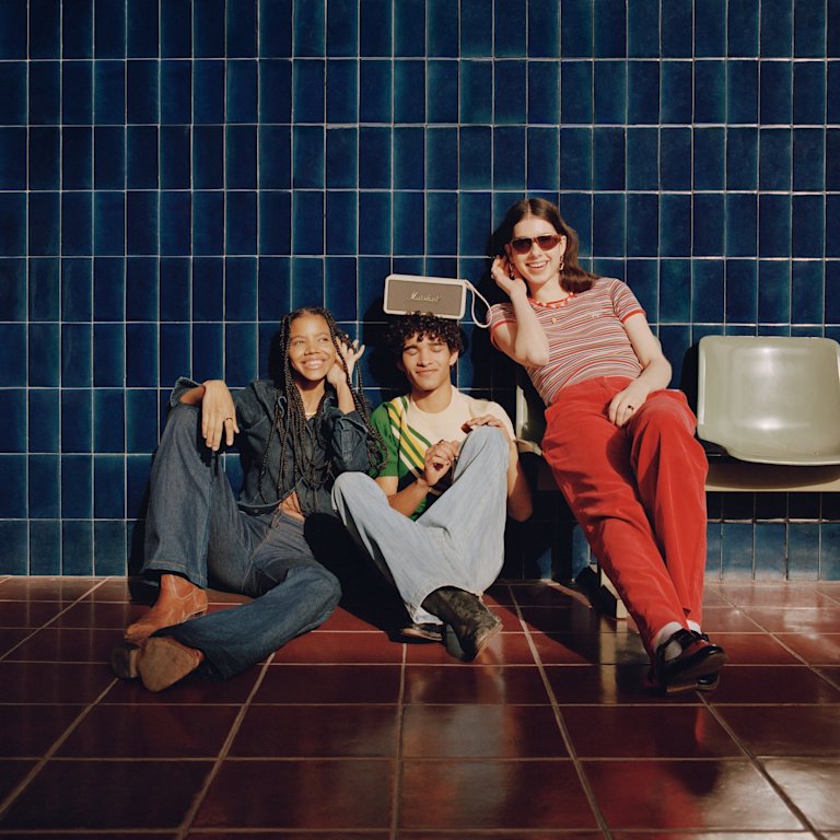 A group of people sitting on the floor in front of a blue tiled wall, with a white Marshall Middleton Cream speaker placed prominently amidst them.