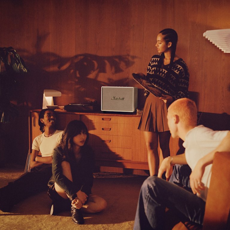 A group of people sitting on the floor in front of a white Marshall WOBURN III CREAM speaker, watching television.