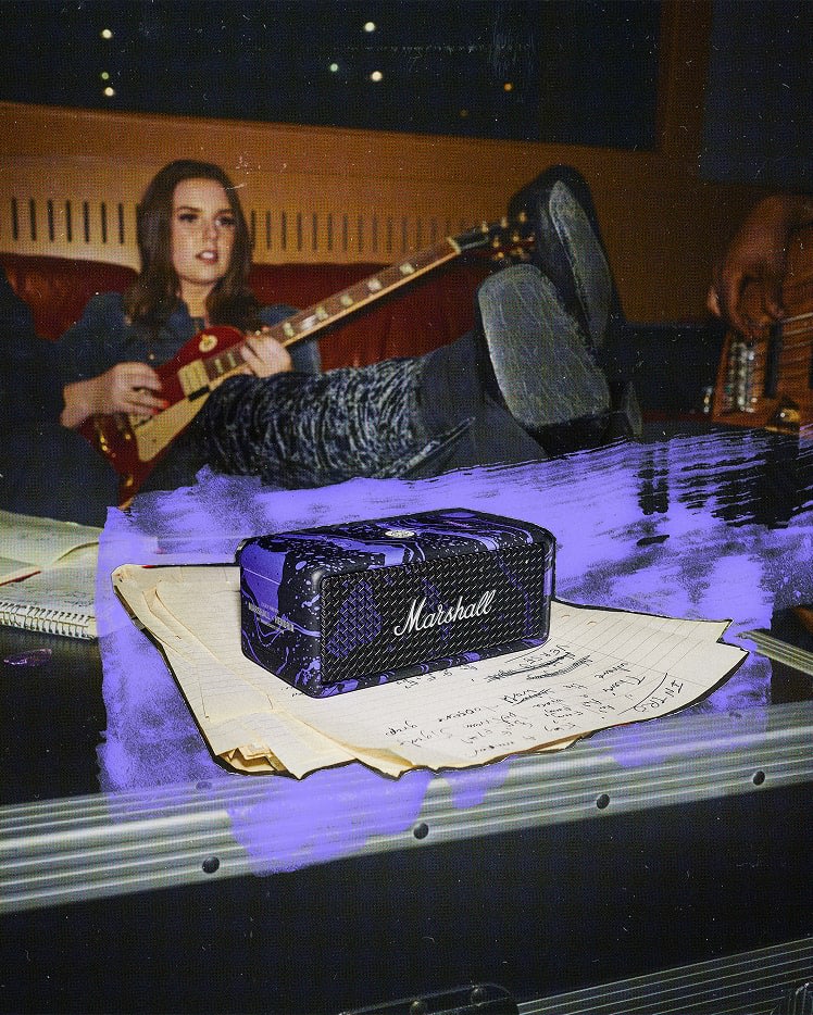 A woman with a guitar sits on a couch, Emberton III Hendrix Edition, papers, and notes are in the foreground.