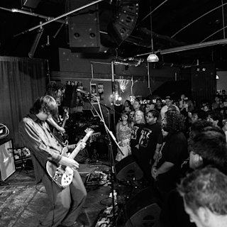 A guitarist performs on stage at an indoor concert venue as a crowd watches. Stage equipment, speakers, and lighting fixtures are visible in the background.