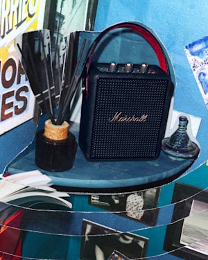 Stockwell III Black and Brass sits on a shelf next to a decorative reed diffuser, a small glass ornament, and assorted photos and posters on a blue wall.