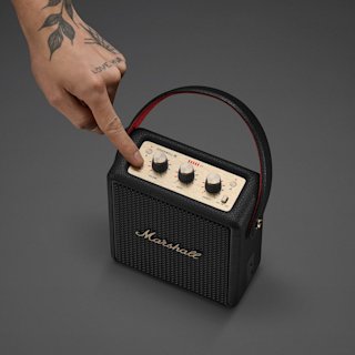 A hand with a tattoo presses the power button on a Marshall Stockwell III Black and Brass speaker.