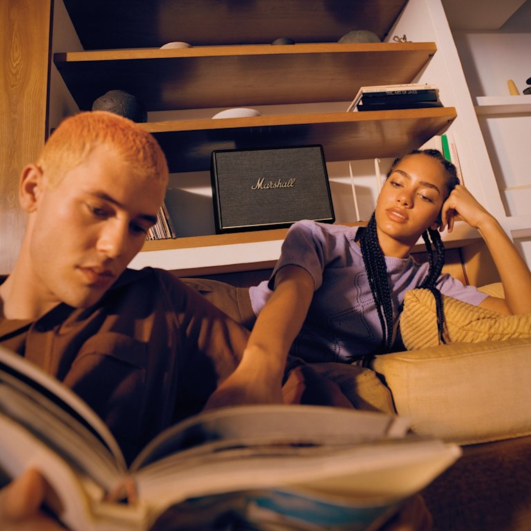 A man and a woman enjoying the Marshall Woburn III Black speaker's exceptional sound quality while reading a book together on their cozy couch.