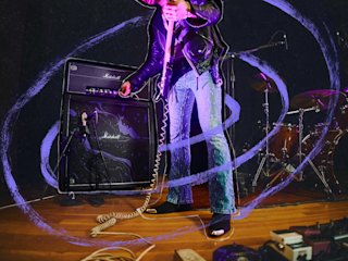 Une personne en jean et veste en cuir règle un amplificateur Marshall x Hendrix 60th Anniversary Collection sur scène, avec une batterie en arrière-plan et des effets lumineux violets tourbillonnants.
