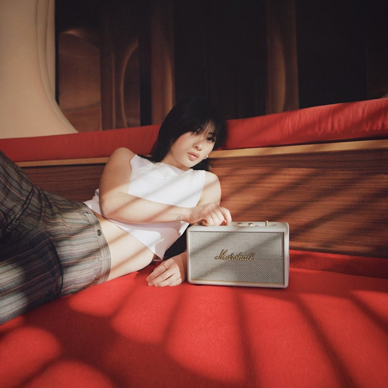 A woman is peacefully lounging on a red couch, accompanied by a Marshall ACTON III Cream speaker.