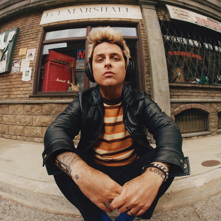 Billie Joe Armstrong wearing Marshall Monitor III A.N.C. Black over-ear headphones, a leather jacket, and a striped shirt sits on the sidewalk.
