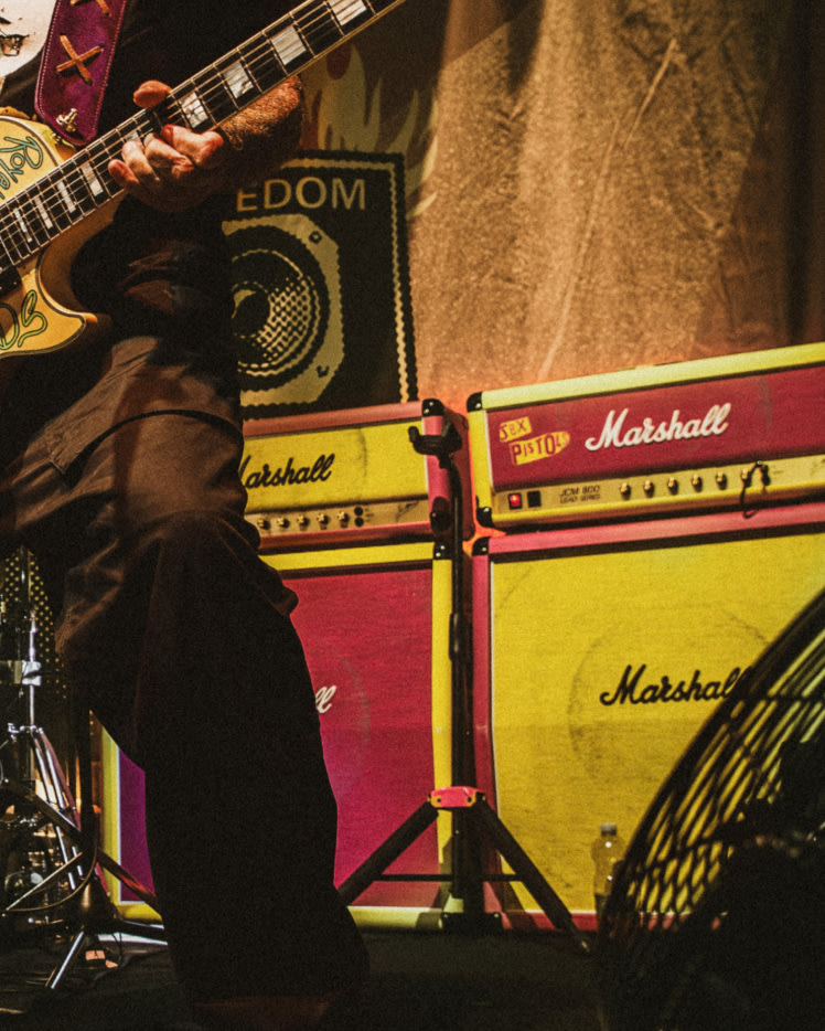 A person is playing a yellow electric guitar with the Sex Pistols amps in the background