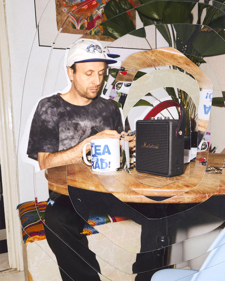 A man sits at a table with a Marshall Stockwell III Black and Brass speakerr, a large mug, and keys.