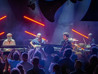 Four musicians perform on stage with keyboards and drums, illuminated by red diagonal lights, while an audience watches and records with smartphones.