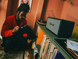 A person in a red sweater holding a record next to a shelf with Stanmore IV Black speaker in a cozy room.