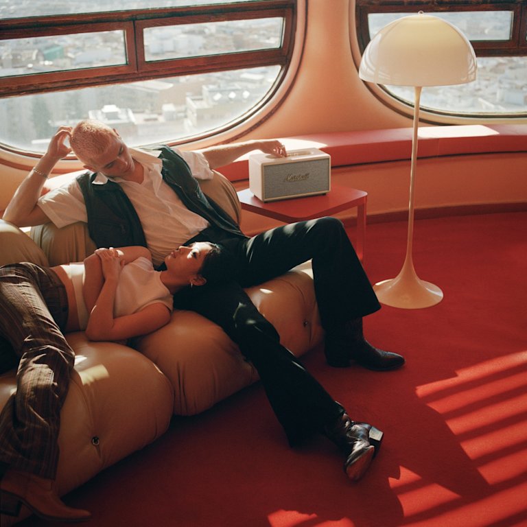 A man and a woman enjoying the comfort of a couch while listening to music on a white Marshall Stanmore III Cream home speaker.