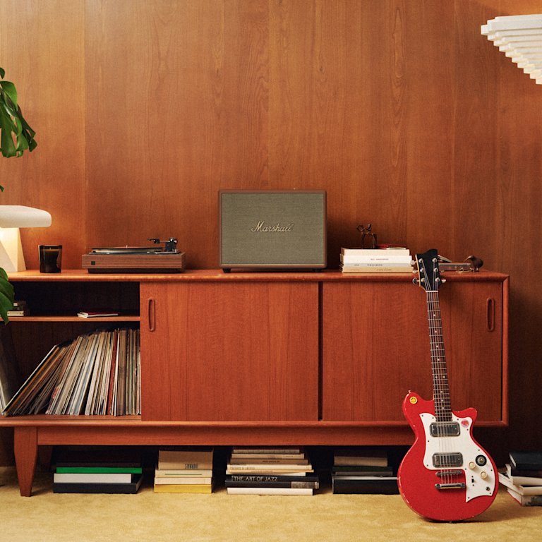 A red electric guitar leans against a wooden cabinet featuring a Marshall Woburn III Brown speaker.