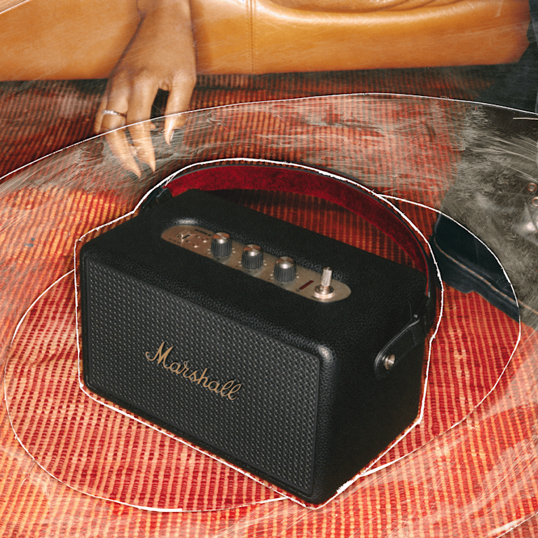 Marshall Kilburn III Black and Brass portable speaker sits on a red patterned rug near a tan leather couch.