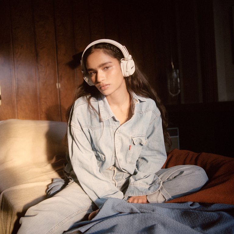 A woman wearing denim clothing and Marshall Major V Cream on-ear headphones sits on a sofa.