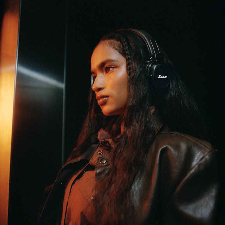A woman with Marshall Major V Brown on-ear headphones and a dark jacket stands in an elevator.
