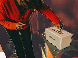A person in a red sweater adjusts the knob on a Stanmore IV Cream speaker placed on a glass table in a room.