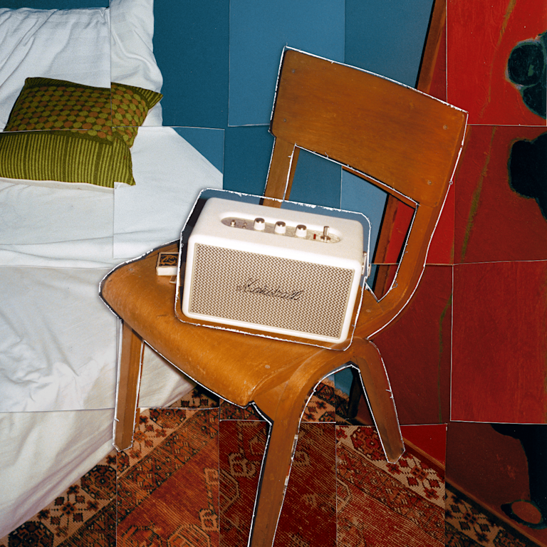 Marshall Kilburn III Cream portable speaker rests on a wooden chair by a bed with white bedding and a green pillow.