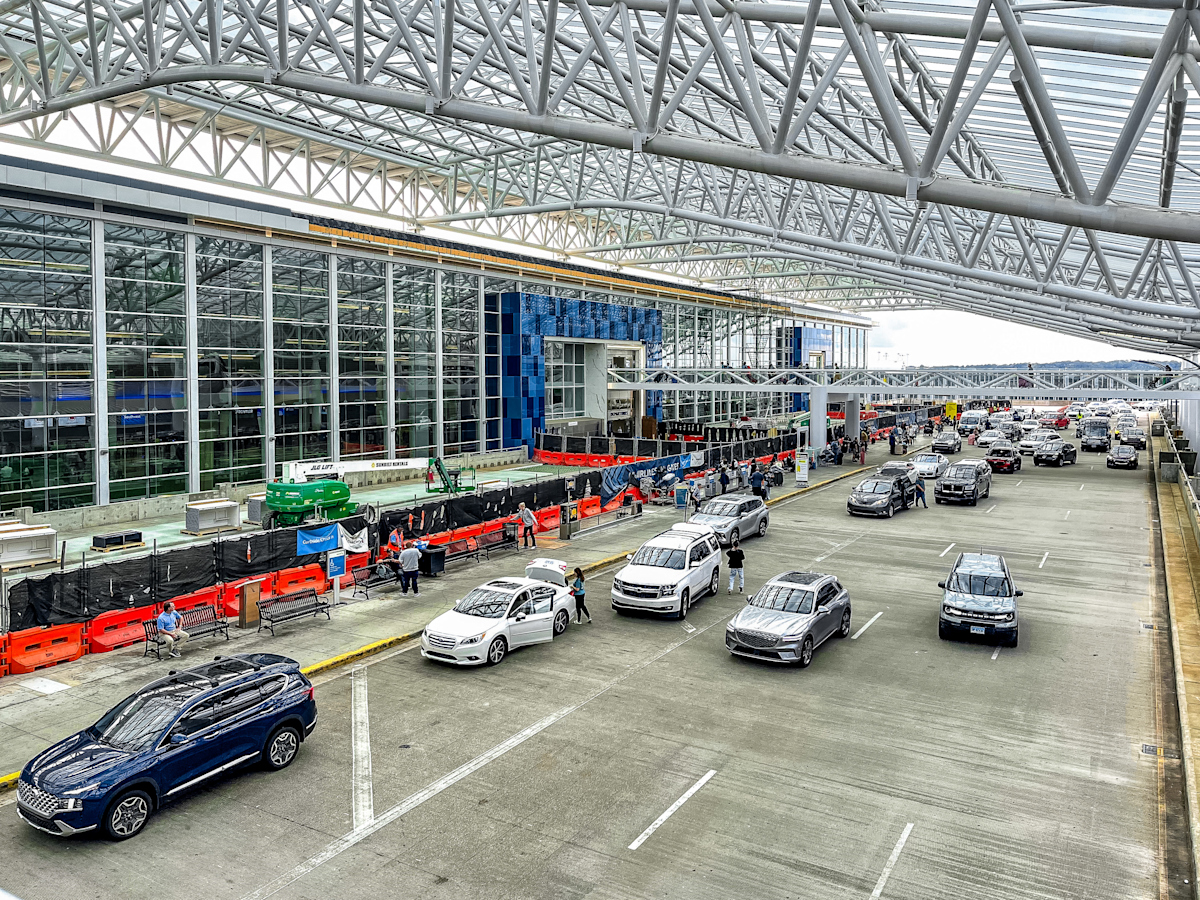 Busy airport drop-off area with cars, passengers, and ongoing terminal construction under a large glass and steel canopy.