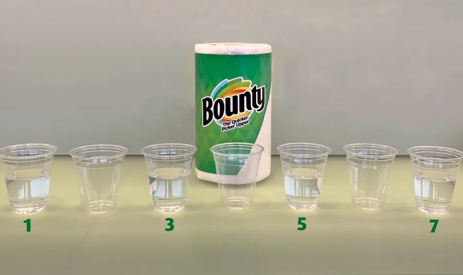 Seven clear cups in front of a Bounty Paper Towel Roll. Every other cup is filled with water