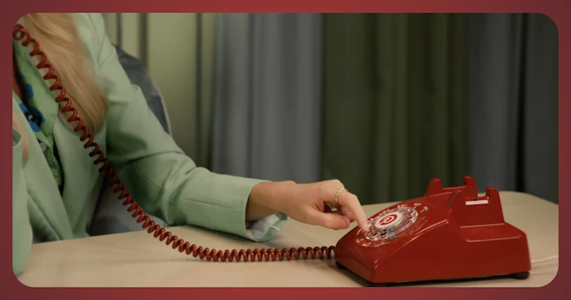 Busto di una donna con la testa fuori dall'inquadratura. È seduta a un tavolo, indossa un completo verde chiaro e compone il numero di un telefono a disco rosso con l'icona "P" di Pinterest al centro.