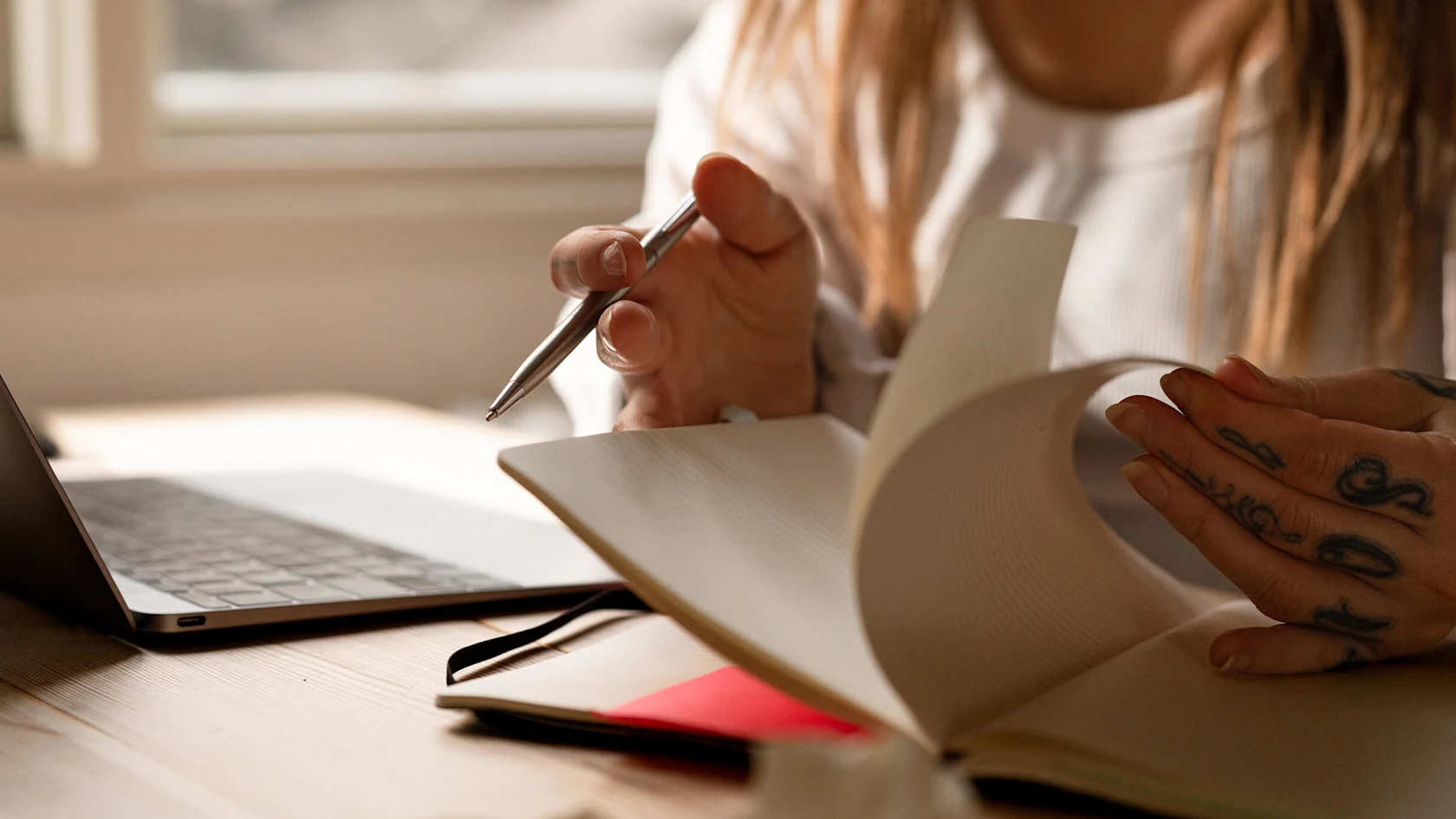 Close-up of a creator’s hands planning in a notebook beside a laptop at a warm, sunlit desk.