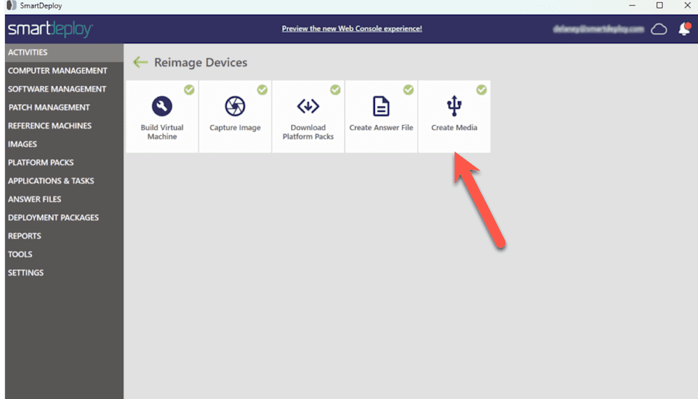 Screenshot of the SmartDeploy interface within the "Reimage Devices" section. A red arrow is pointing to the "Create Media" tile, which features a USB icon and is the last option in a row of five deployment tasks. The other tiles include "Build Virtual Machine," "Capture Image," "Download Platform Packs," and "Create Answer File." Each tile has a green checkmark indicating completion or availability. The left-side navigation menu lists categories such as Activities, Images, Platform Packs, and more. The header includes a link to preview the new Web Console experience and displays the logged-in user's email.