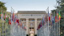 Bild vom Gebäude der Vereinten Nationen in der Stadt Genf, Schweiz. Die Vorderseite des Gebäudes ist mit vielen Fahnen und Fahnenmasten gesäumt, die alle Mitgliedsnationen der UNO repräsentieren.
