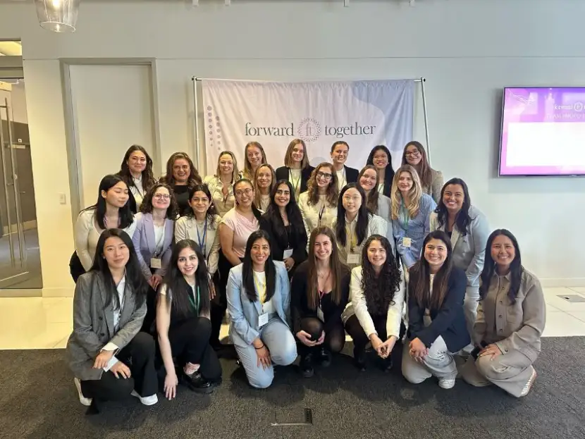 Un grand groupe de femmes pose ensemble en souriant devant une bannière affichant « forward together » lors d’un événement intérieur.