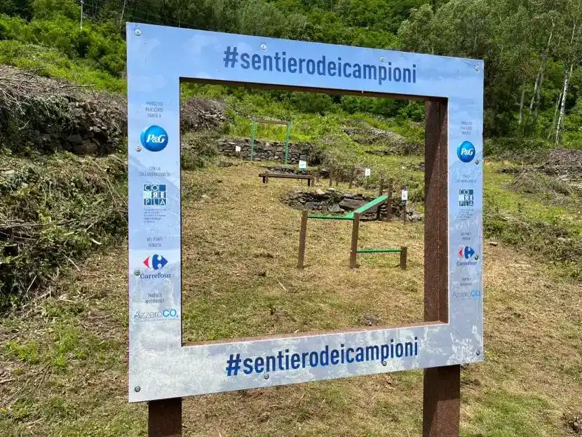 na cornice azzurra con la scritta sentiero dei campioni, loghi di P&G carrefour, corepla, azzeroco2. Attraverso la cornice si vede una delle 5 zone ti attività del percorso con attrezzature sportive