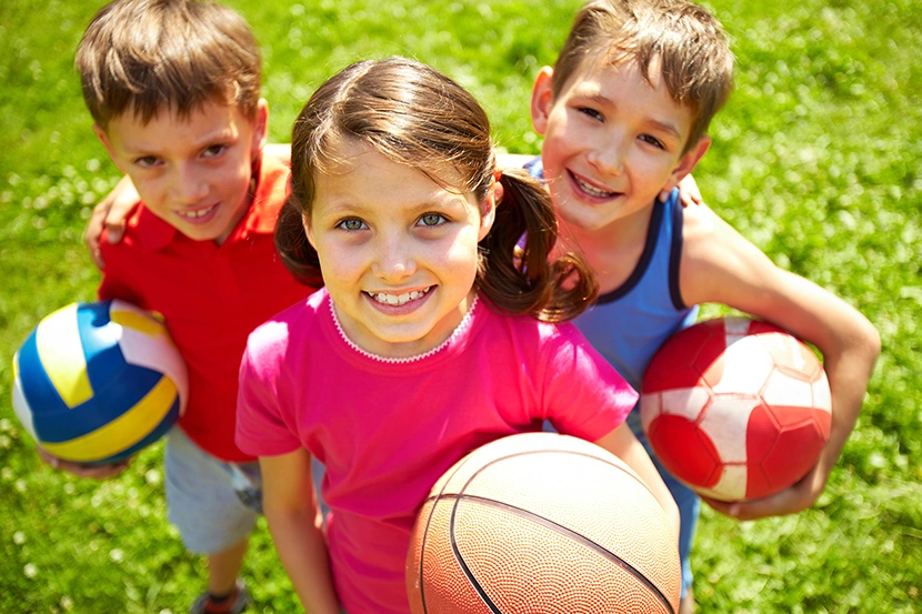 Tre bambini sorridenti con in mano palloni sportivi mentre stanno insieme su un prato.