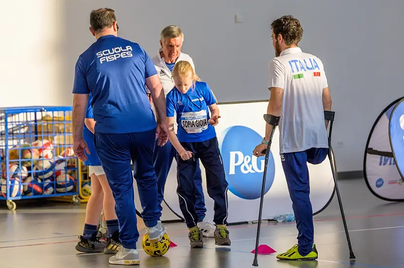 Un ragazzo con una gamba amputata si regge sulle stampelle mentre parla con una bambina con sindrome di autismo, sorretta da un insegnante di ginnastica. Si trovano in una palestra e stanno giocando a pallone