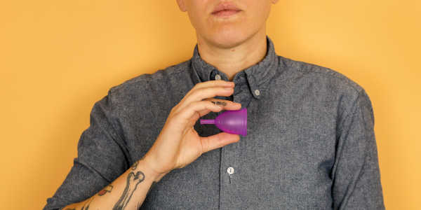 一个手臂上有纹身，身穿灰色衬衫的人拿着一个紫色的红宝石杯月经杯. 