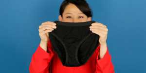 A woman with black hair and a red shirt holds a pair of black Ooshi bikini-style underwear.