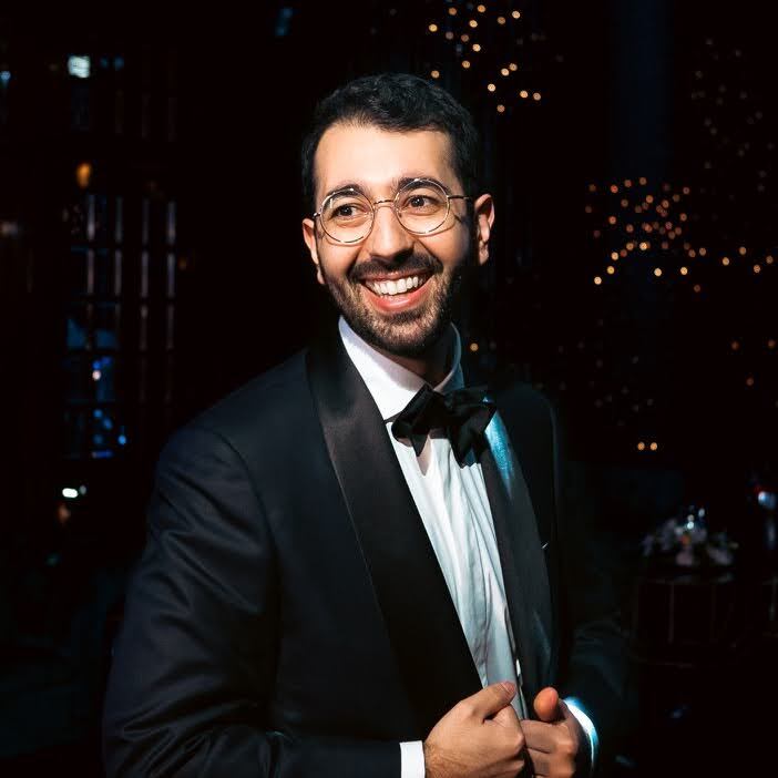 Person in formal black tuxedo and bow tie smiling at a nighttime event with twinkling lights in background.