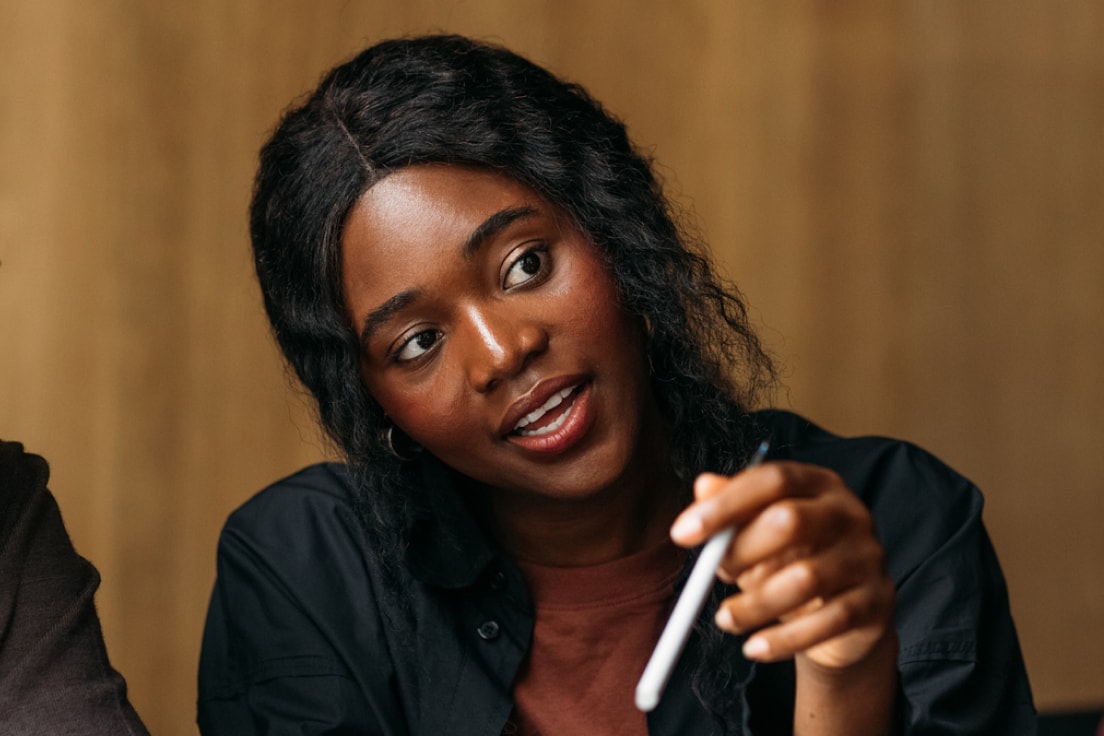 Person with dark curly hair in black shirt gesturing with pen during conversation against wooden background.