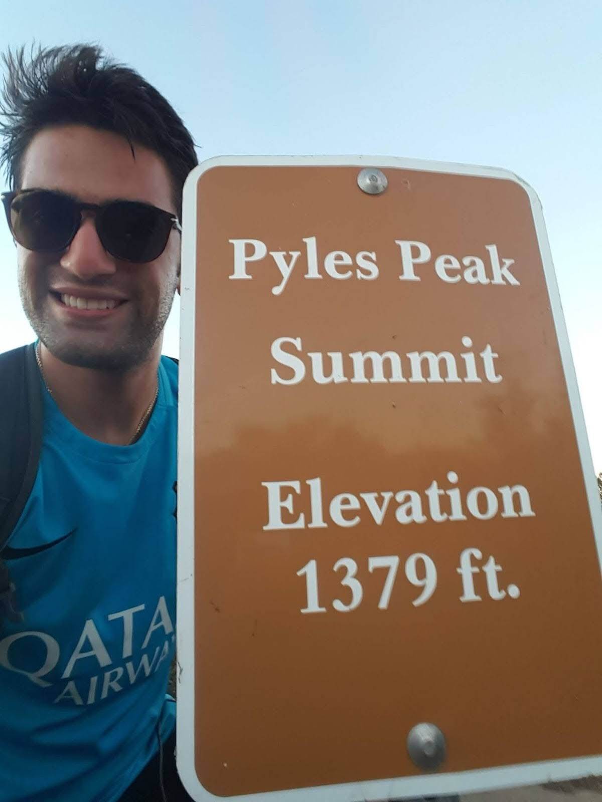 Person in blue shirt and sunglasses next to Pyles Peak Summit sign showing elevation of 1379 ft.