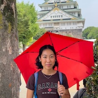 Rippler Sheline Sim standing among trees, with impressive architecture in the background, while holding a red umbrella.