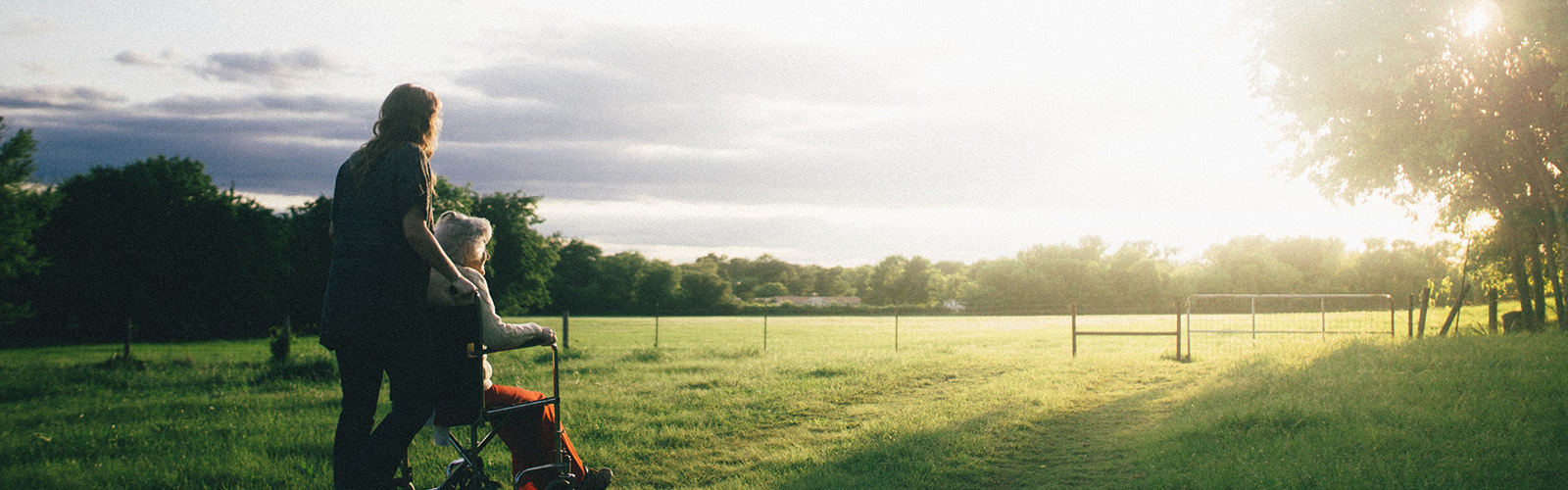 A woman pushing an older woman in a wheelchair through a field at dawn with the sun rising in the horizon