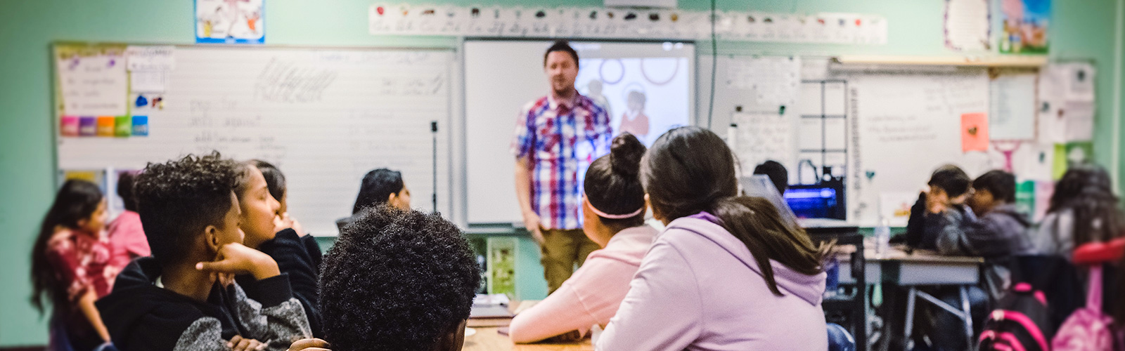 A class of students sat at their tables with a teacher at the front teaching