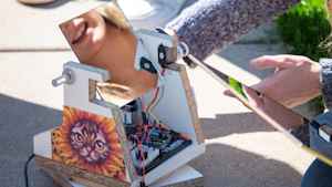 A female student's smile is reflected in the mirror of a solar powered camera built from wires and particle board by her and her classmates. On the machine is taped a printout of a cat wearing a sunflower costume.