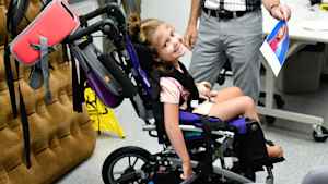 A young girl sits in an oversized stroller and smiles over her shoulder. The occupational therapist shows her a paper printout of the two princesses from the movie